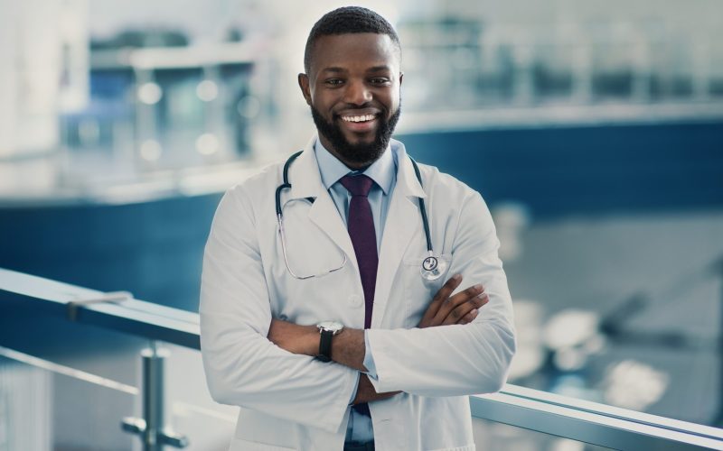 Portrait of handsome smiling young african american doctor in workwear and stethoscope with arms crossed on chest posing at clinic hall, copy space. Modern healtcare, hospitals and clinics concept